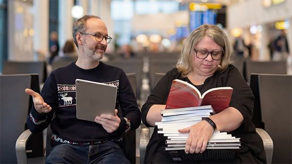 Deux personnes assises côte à côte : l'une tient une tablette numérique et l'autre une pile de livres.